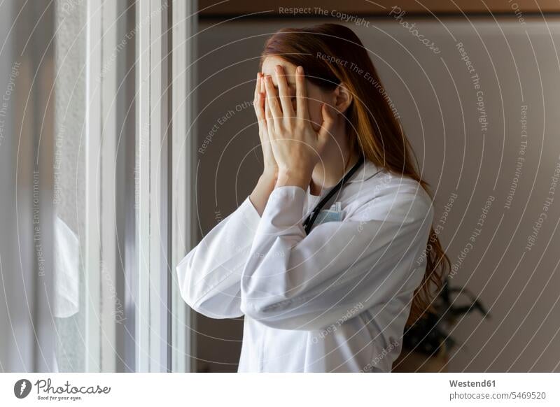 Emotionally stressed young female doctor with head in hands standing by window color image colour image Female Doctor Female Doctors doctors physician