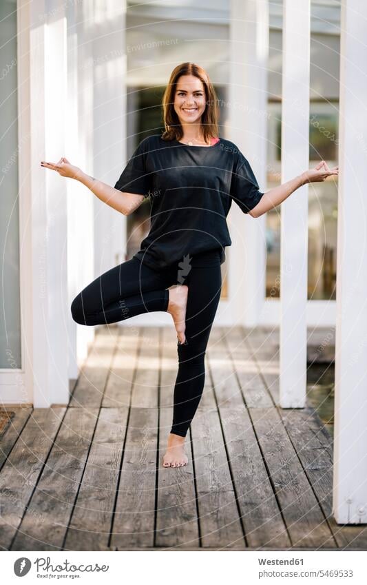 Smiling mid adult woman practicing tree pose on hardwood floor against house color image colour image outdoors location shots outdoor shot outdoor shots day