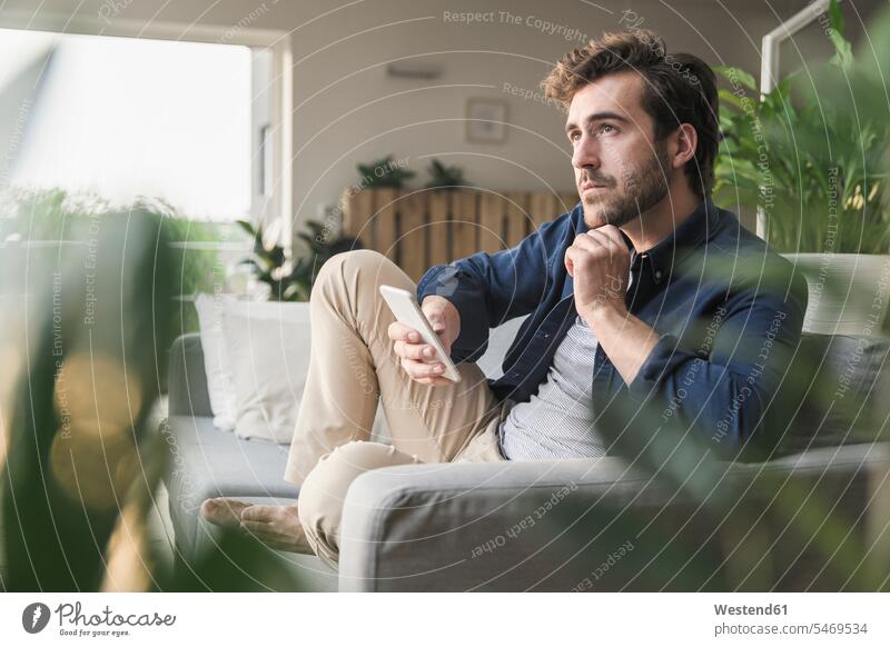 Young man sitting on couch at home, using smartphone settee sofa sofas couches settees Contemplation reflection Contemplating contemplate thinking independence