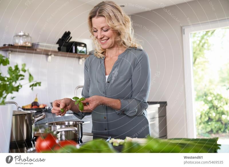 Smiling woman cooking in kitchen putting basil into cooking pot Basils smiling smile Cooking Pot Pots Cooking Pots females women domestic kitchen kitchens