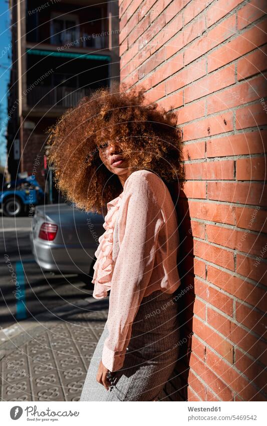 Portrait of beautiful young woman with afro hairdo leaning against brick wall in the city town cities towns females women Afro portrait portraits outdoors