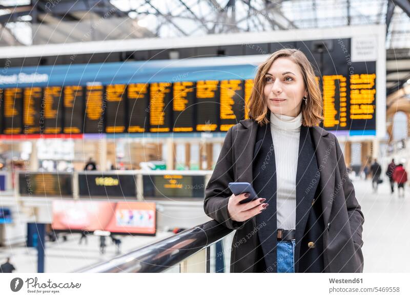 Woman at train station holding a mobile phone, London, UK coat coats jackets telecommunication phones telephone telephones cell phone cell phones Cellphone