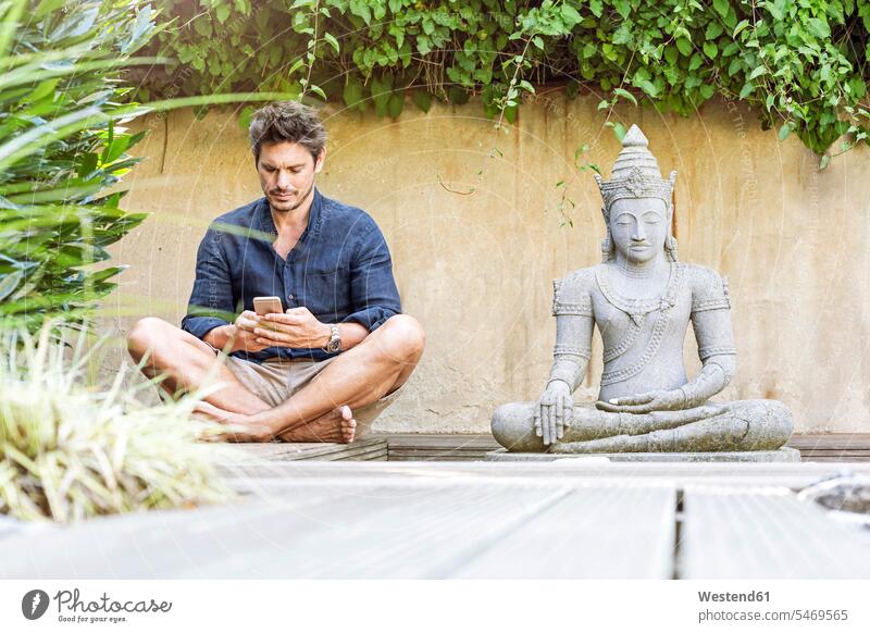 Man sitting cross-legged next to Buddha statue in a Zen garden, using smartphone mobile phone mobiles mobile phones Cellphone cell phone cell phones