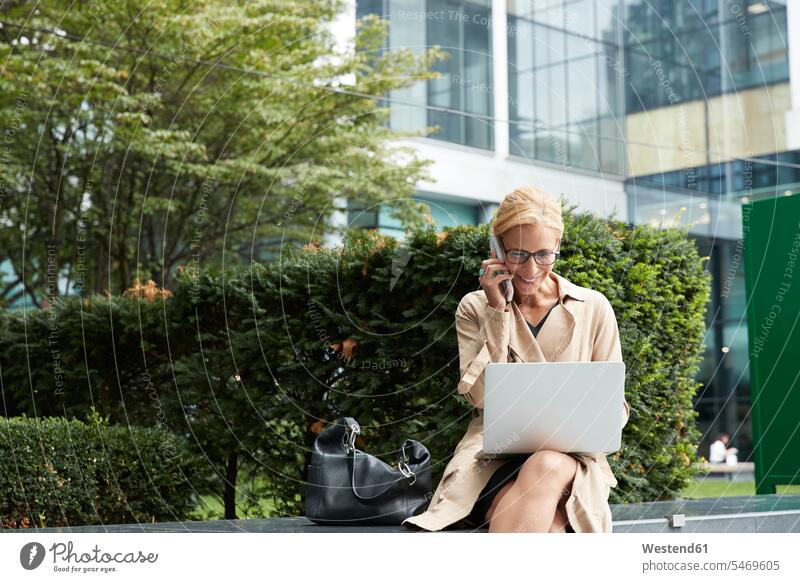 Businesswoman talking on mobile phone while working on laptop at office park color image colour image outdoors location shots outdoor shot outdoor shots day