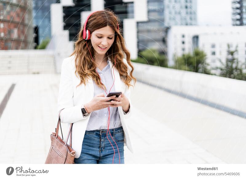 Young businesswoman commuting in the city, using smartphone and headphones on the move on the way on the go on the road urban urbanity smiling smile use walking