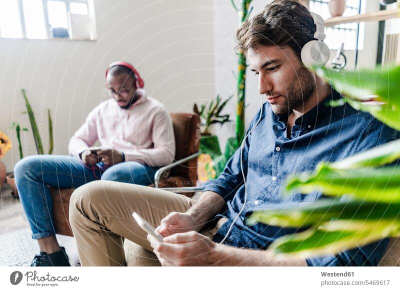 Two young men with cell phones and headphones sitting in armchairs in a loft mobile phone mobiles mobile phones Cellphone lofts man males headset Arm Chairs