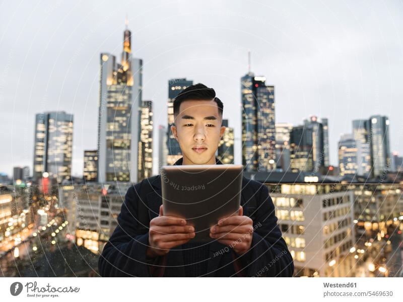 Man using tablet in front of urban skyline at dusk, Frankfurt, Germany human human being human beings humans person persons Asian Asians Japanese 1