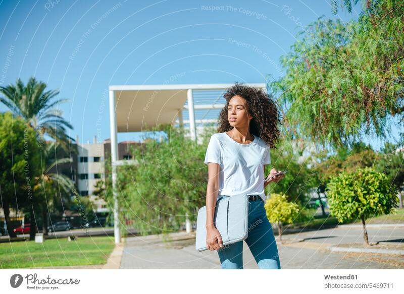 Portrait of young woman with mobile phone and laptop bag walking down the street looking at cell phone laptop case females women going view seeing viewing