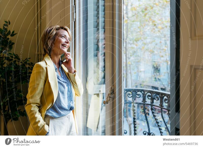 Smiling businesswoman talking on smart phone while looking through window standing in office color image colour image indoors indoor shot indoor shots interior
