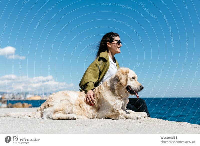 Happy woman sitting besides her Labrador Retriever at quay looking to the sea dog owner horizon copy space horizons walking going go walkies walking the dogs