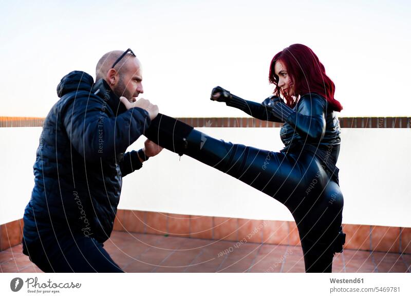 Redheaded woman wearing black leather catsuit fighting against man on rooftop gloves step Tread Treading colour colours stand combative sport Power Spiritedness