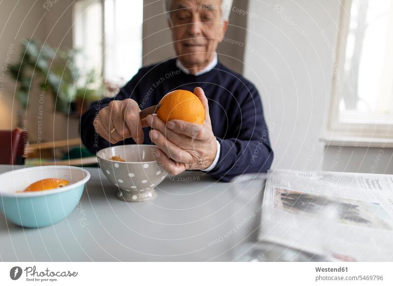 Senior man peeling orange at home Bowls windows newspapers jumper sweater Sweaters Tables hold cut Seated sit in the morning Everyday Everyday Scene