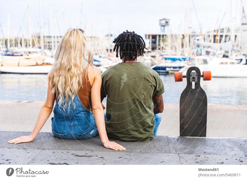 Spain, Barcelona, back view of multicultural young couple sitting side by side with longboard at marina Seated Longboard twosomes partnership couples paralell