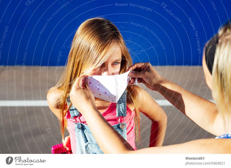 Mother putting protective mask on daughter's face during sunny day color image colour image outdoors location shots outdoor shot outdoor shots daylight shot