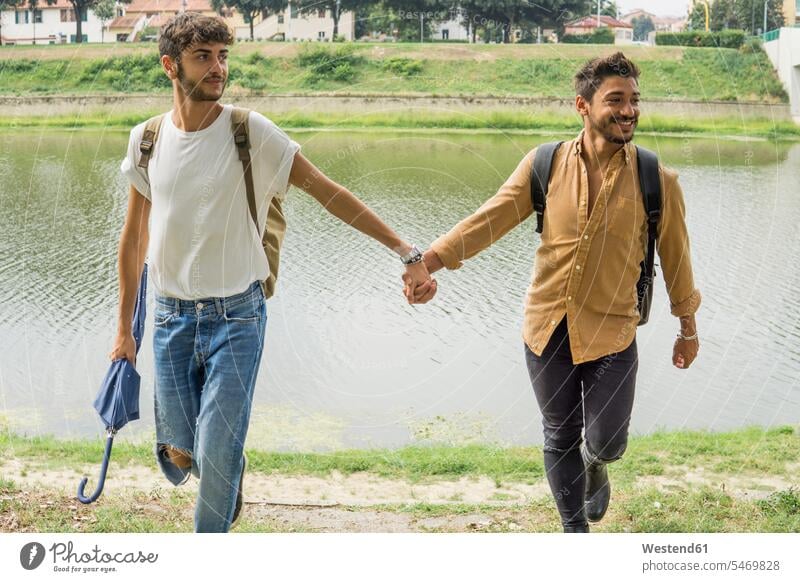 Portrait of young gay couple with backpacks walking hand in hand at riverside riverbank rucksacks back-packs going gay men gay man homosexual men homosexual man