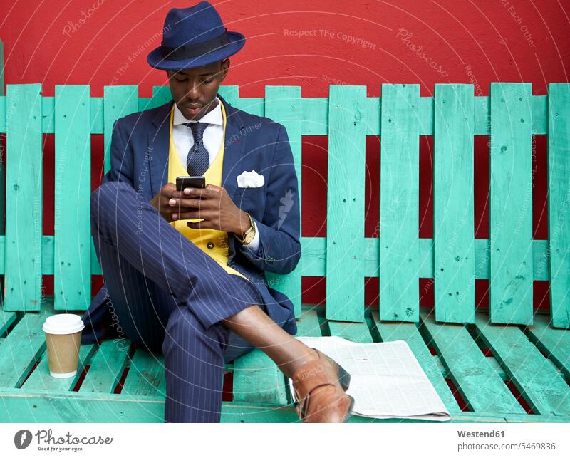Young businessman wearing old-fashioned suit and hat sitting on a green bench checking his phone business life business world business person businesspeople