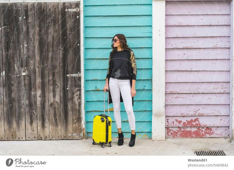 Young woman with a yellow trolley bag waiting in front of wooden facade Trolley Bag females women colour colours rolling suitcase rolling suitcases Adults