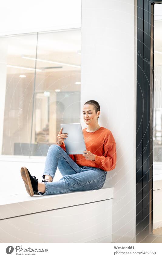 Relaxed businesswoman sitting on windowsill in office building, using digital tablet human human being human beings humans person persons caucasian appearance