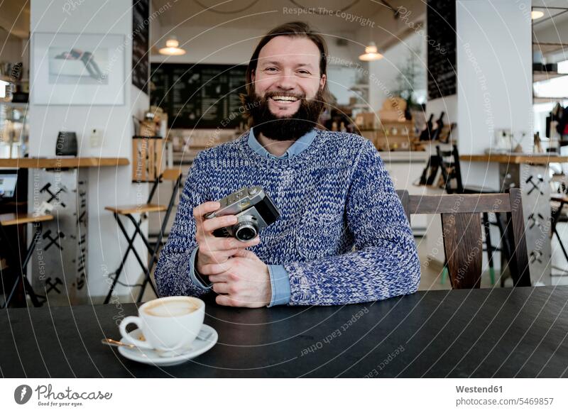 Man with beard sitting in cafe, holding old camera man men males cameras photographer photographers full beard drinking laughing Laughter smiling smile amiable