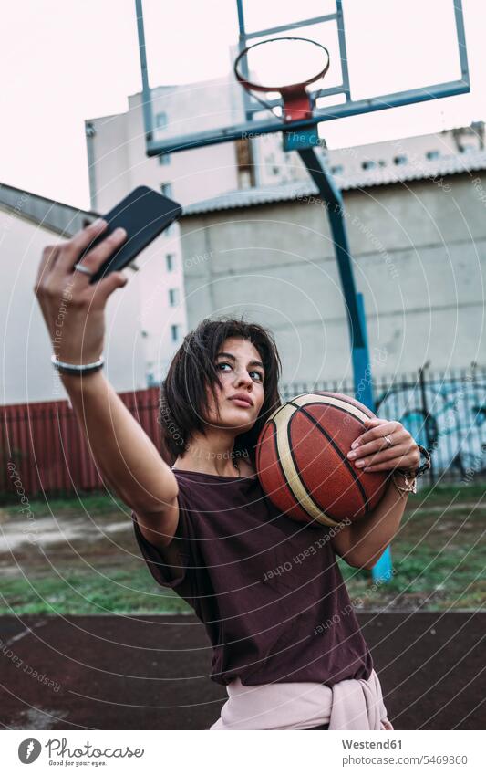 Young woman with basketball taking a selfie on outdoor court basketballs Selfie Selfies sports field sports fields females women Basketball Adults grown-ups