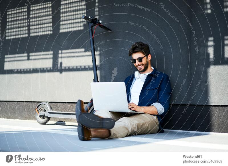 Smiling young man sitting on the ground besides his electric scooter using laptop Seated use land floor men males next to E-Scooter Laptop Computers laptops