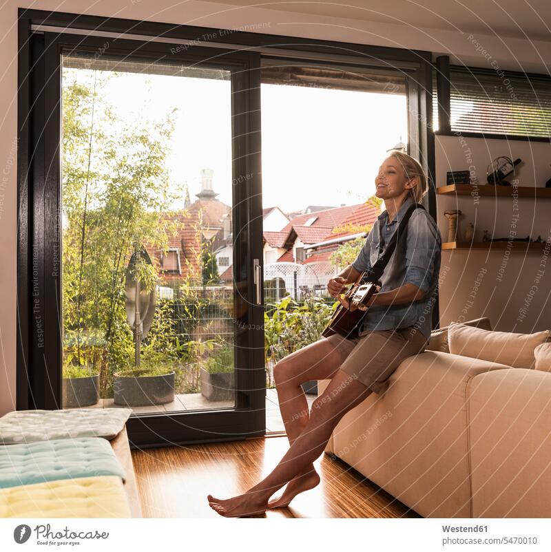 Mature woman singing and playing guitar in the living room females women living rooms livingroom guitars Adults grown-ups grownups adult people persons