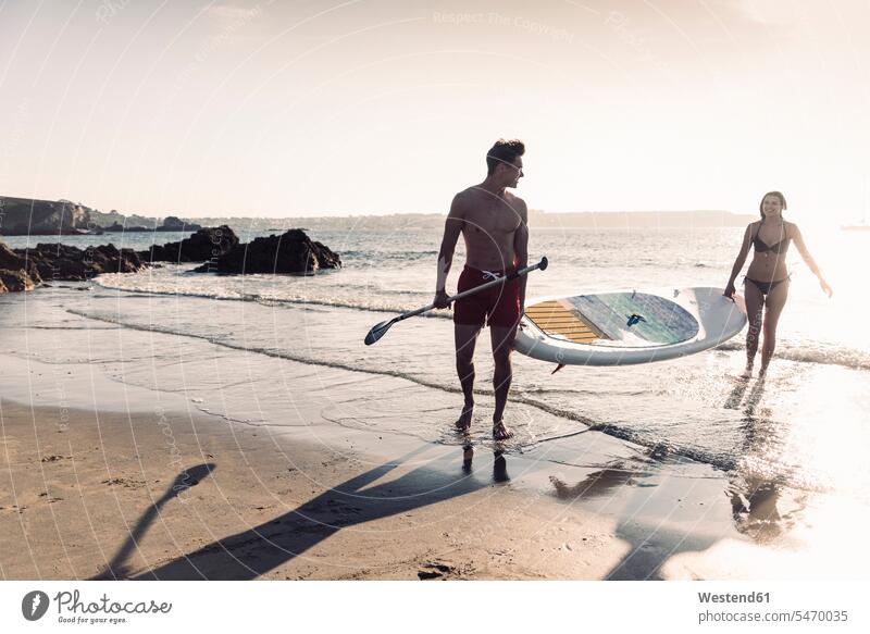 France, Brittany, young couple carrying an SUP board at the sea together ocean Paddleboard standup paddleboard paddle board Paddleboards twosomes partnership