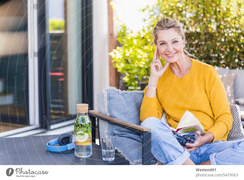 Portrait of smiling mature woman sitting on terrace with bowl of blueberries and book Bottles Glass Bottles Crockery Tableware Drinking Glass Drinking Glasses