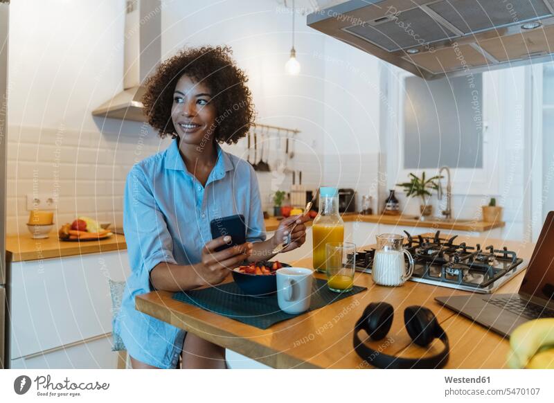 Woman having breakfast in her kitchen, using smartphone use smiling smile laptop Laptop Computers laptops notebook domestic kitchen kitchens sitting Seated