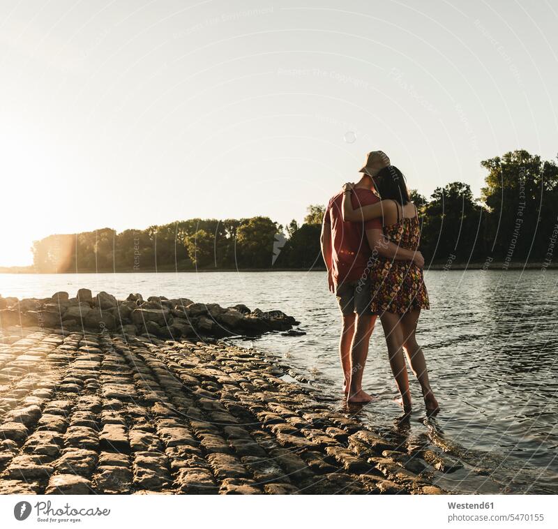 Rear view of young couple embracing at a river at sunset sunsets sundown embrace Embracement hug hugging River Rivers twosomes partnership couples atmosphere