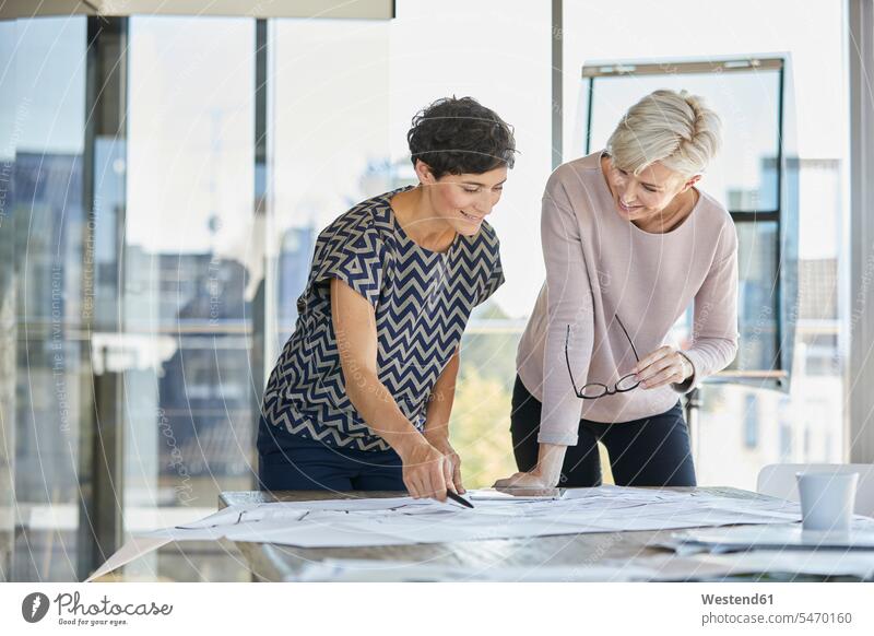 Two smiling businesswomen discussing plan on desk in office businesswoman business woman business women offices office room office rooms plans working At Work