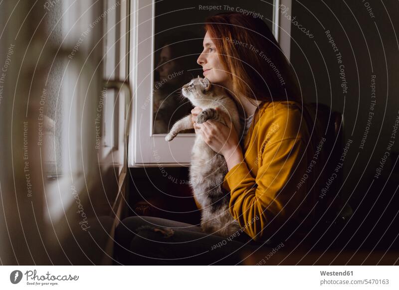 Smiling redheaded woman sitting with her cat at open window at home human human being human beings humans person persons celibate celibates singles