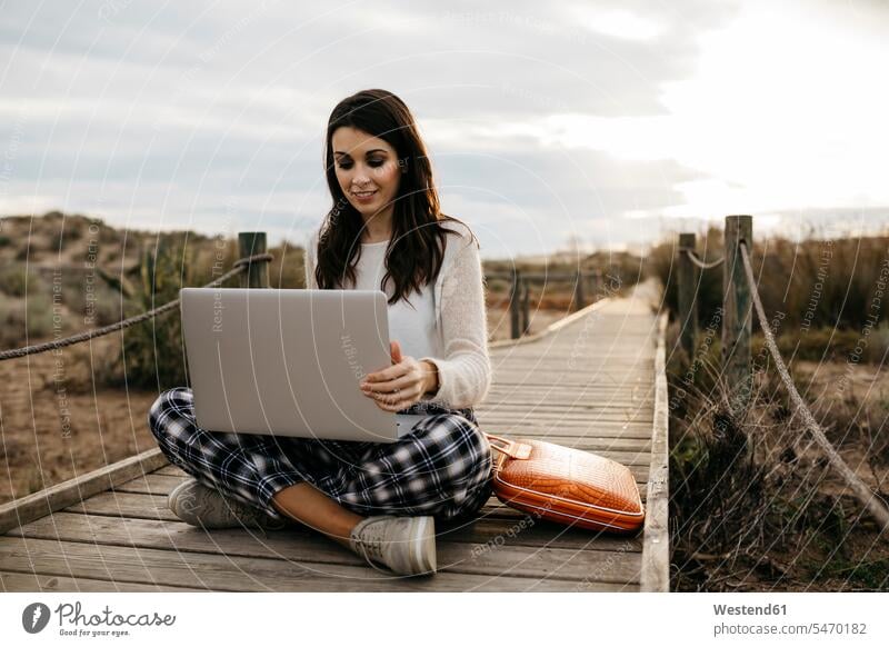 Woman sitting on a boardwalk in the countryside using laptop bags computers Laptop Computer Laptop Computers laptops notebook At Work work smile Seated solitary