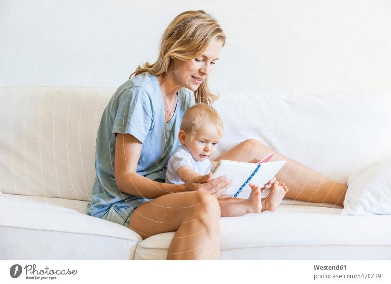 Mother and baby girl sitting on couch looking at picture book infants nurselings babies eyeing Picture Books picture-books settee sofa sofas couches settees