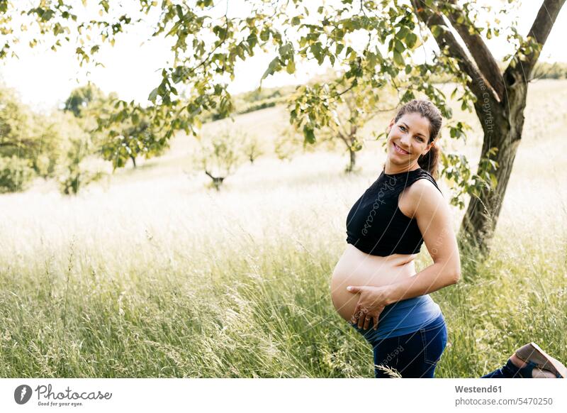 Young pregnant woman holding baby belly, standing on a meadow upper part of the body bellies stomach stomachs touch smile delight enjoyment Pleasant pleasure