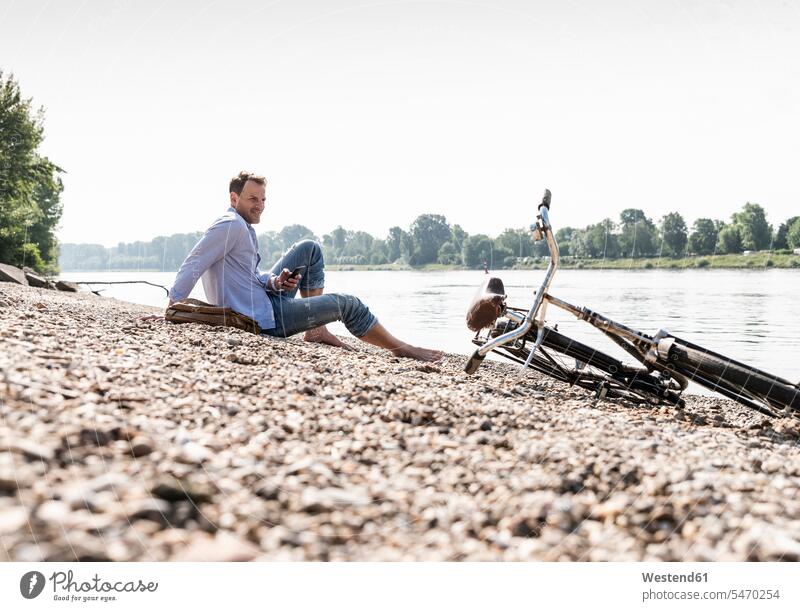 Mature man with bike and smartphone sitting at Rhine riverbank men males bicycle bikes bicycles Seated water's edge waterside shore cyclist Smartphone iPhone