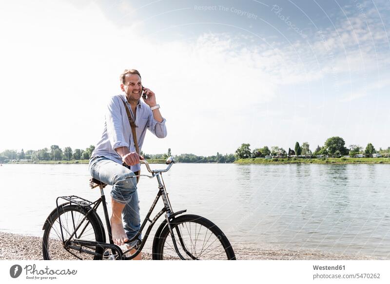 Mature man with bike using smartphone at Rhine riverbank bicycle bikes bicycles use cyclist Smartphone iPhone Smartphones call telephone call Phone Call
