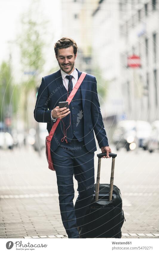 Smiling businessman walking in the city with cell phone and suitcase Germany Smartphone iPhone Smartphones Traveller Travellers Travelers freelancer freelancing
