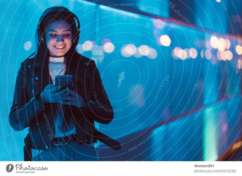 Portrait of smiling teenage girl with headphones standing in front of blue glass pane looking at smartphone human human being human beings humans person persons