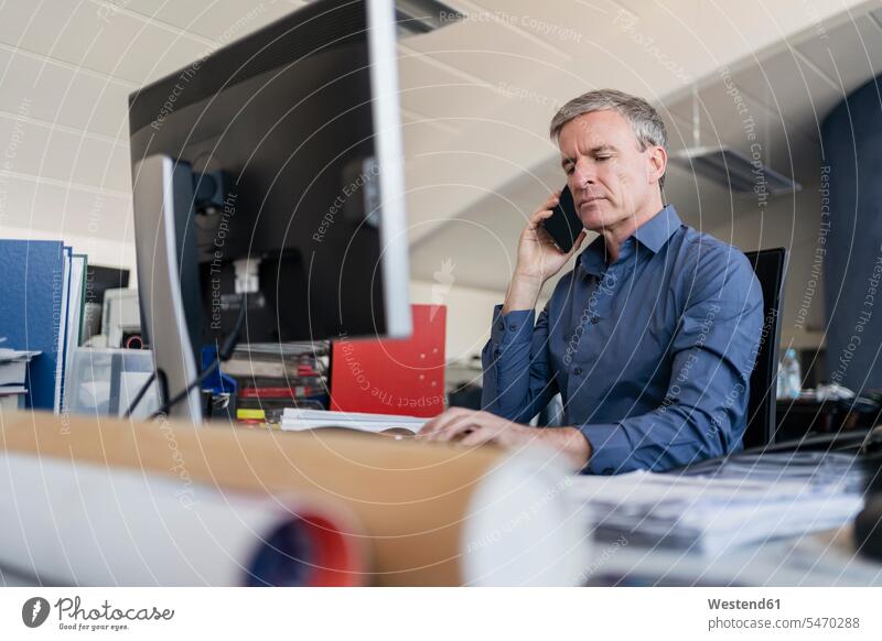 Handsome male professional talking on mobile phone while sitting at computer desk in office color image colour image Germany indoors indoor shot indoor shots