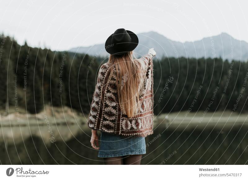 Back view of fashionable young woman wearing hat and poncho standing in front of a lake hats lakes females women water waters body of water Adults grown-ups