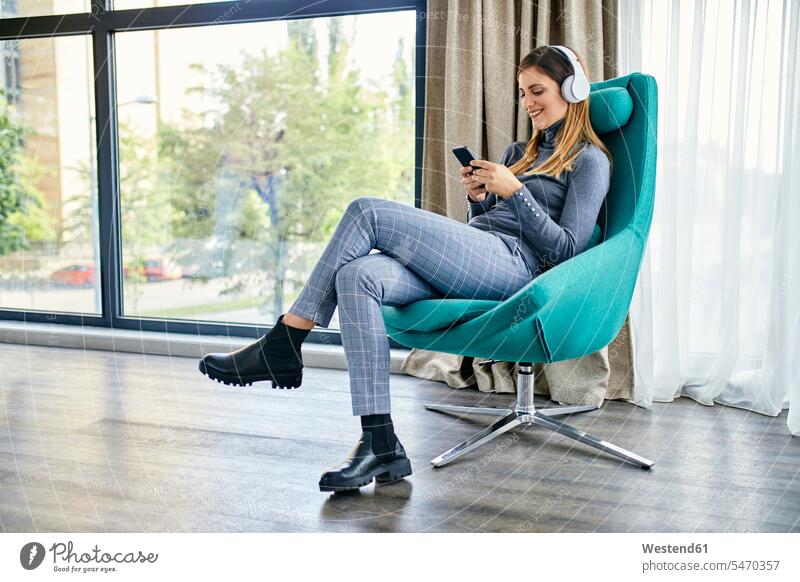 Woman sitting in armchair, listening music with headphones, using smartphone human human being human beings humans person persons caucasian appearance