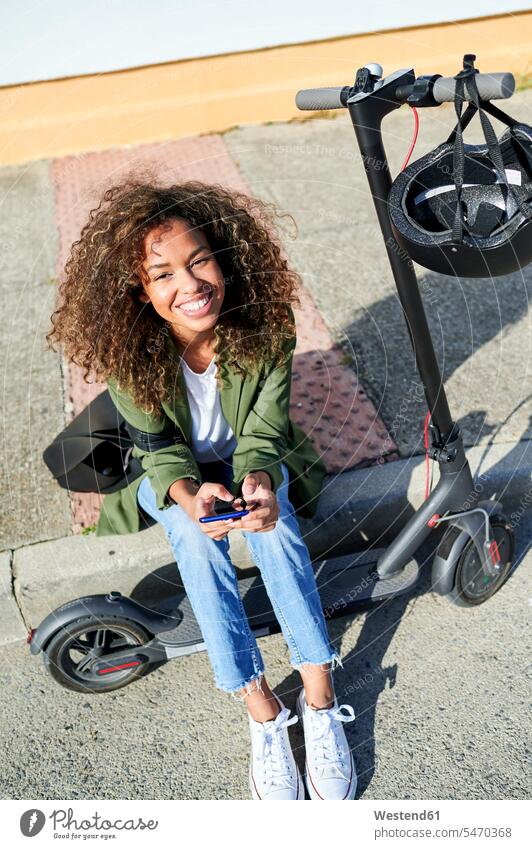 Happy young woman holding smart phone while sitting on sidewalk with electric push scooter color image colour image outdoors location shots outdoor shot