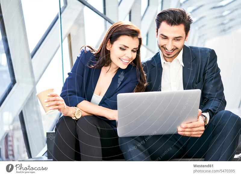 Smiling businesswoman and businessman sharing laptop on stairs in modern office Laptop Computers laptops notebook sitting Seated smiling smile Businessman