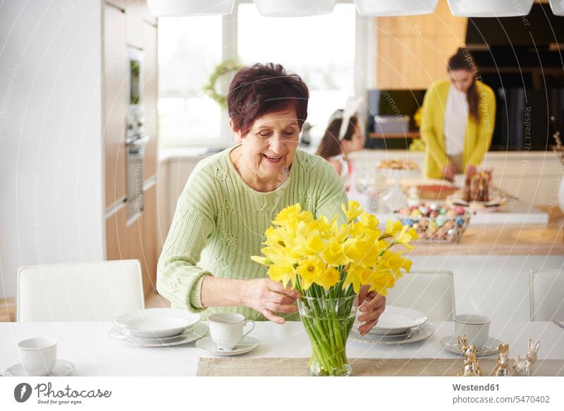 Senior woman arranging flowers on dining table with family in background girl females girls Flower Flowers families home at home Dining Table Dinner Table