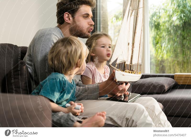 Father and children blowing into sails of toy model ship on couch at home settee sofa sofas couches settees father pa fathers daddy dads papa model boat family