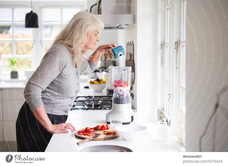 Senior woman pouring milk in processor while preparing strawberry smoothie at kitchen color image colour image Denmark Scandinavia Scandinavian Peninsula