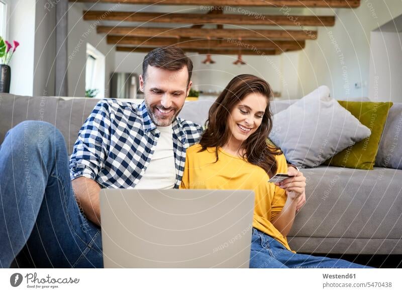 Couple sitting in living room, using laptop, making an online payment with their credit card debit card using a laptop Using Laptops living rooms livingroom
