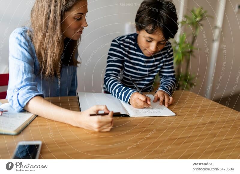 Female tutor looking at boy writing homework in book on table color image colour image Spain casual clothing casual wear leisure wear casual clothes