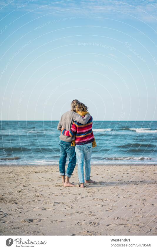 ture couple standing on the beach with arms around, looking at the sea ocean beaches twosomes partnership couples watching water waters body of water people